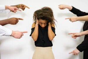 Woman covering her ears with her face down in a stressed manner, while 7 hands are all pointing at her causing conflict.